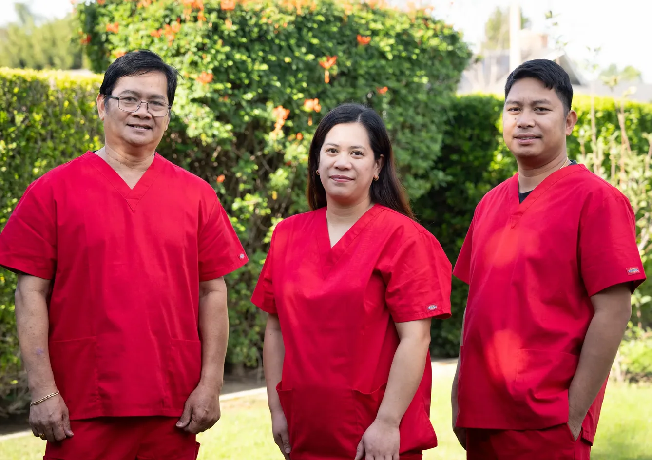 Three professional caregivers in red medical scrubs posing together in the facility's outdoor garden area